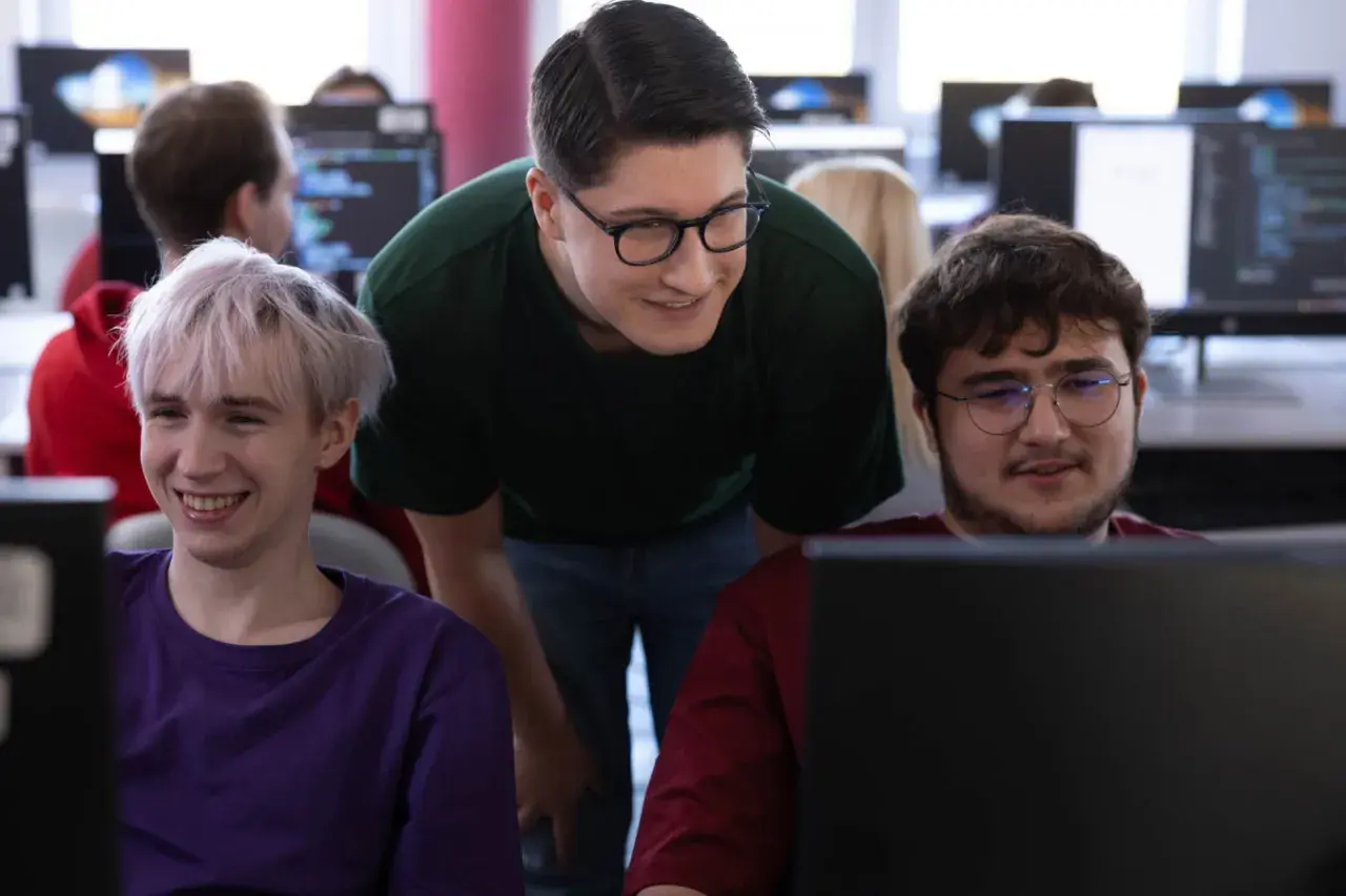 Trzech młodych informatyków przy komputerach. Jeden z nich, w zielonej koszulce, pochyla się nad kolegami, dyskutując o kodzie. Zastanawiasz się, ile zarabia informatyk po studiach?